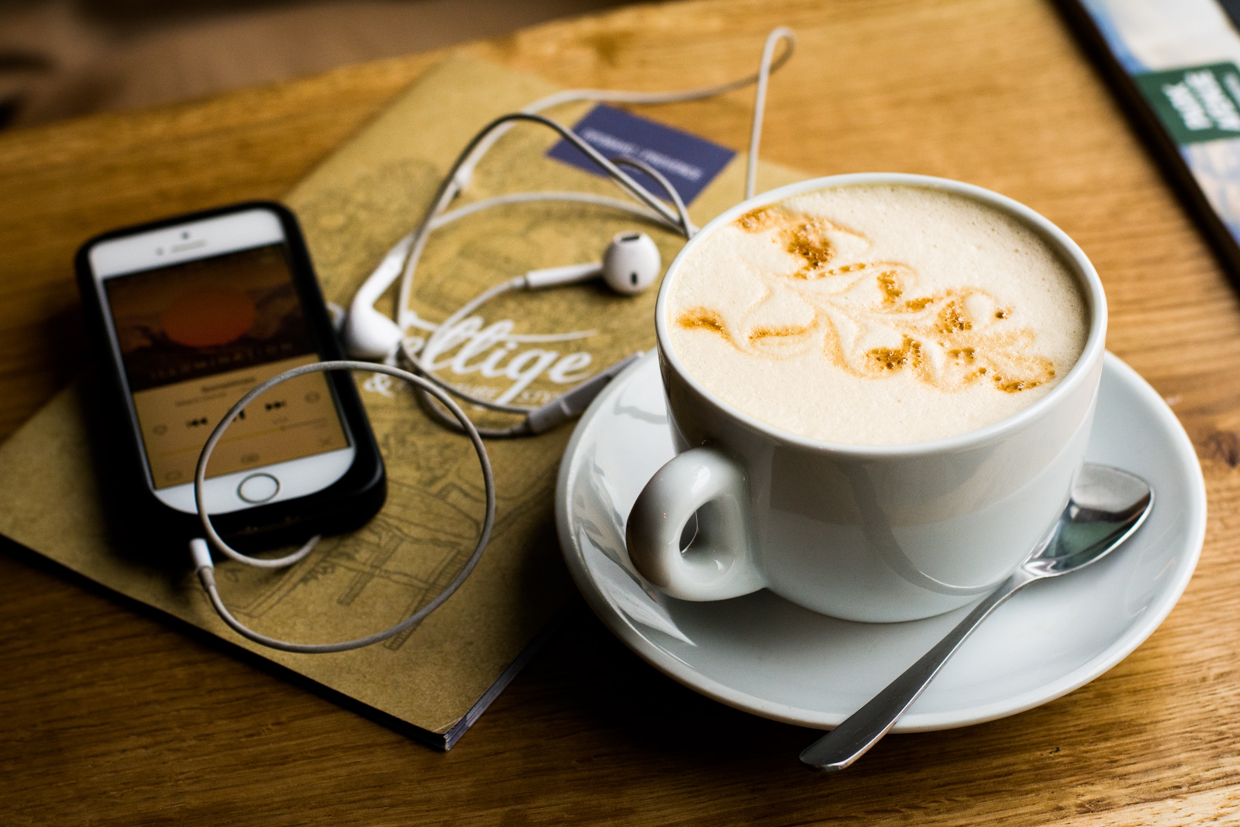An iphone playing a podcast with headphones plugged in. On a table next to a cappuccino. 
